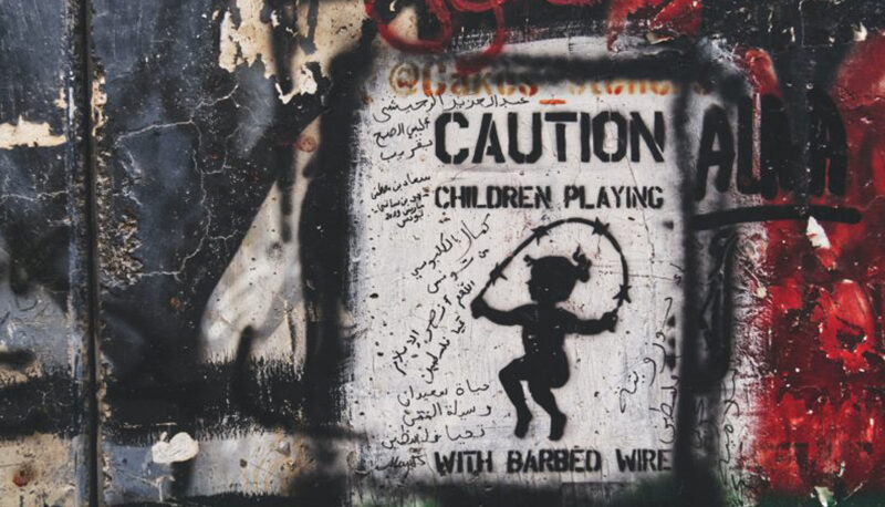 Artwork on the wall that separating Israel and Palestine. The main art is a black stencil of a child jump roping with barbed wire, with the words, "CAUTION: CHILDREN PLAYING WITH BARBED WIRE."