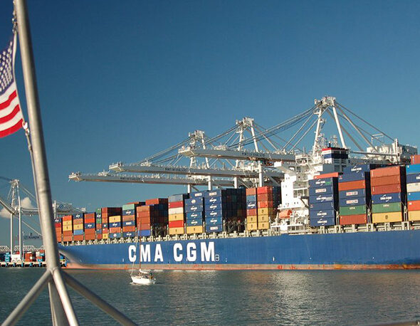 An image of a cargo ship transporting multiple colorful shipping containers. An American flag waves in the foreground.