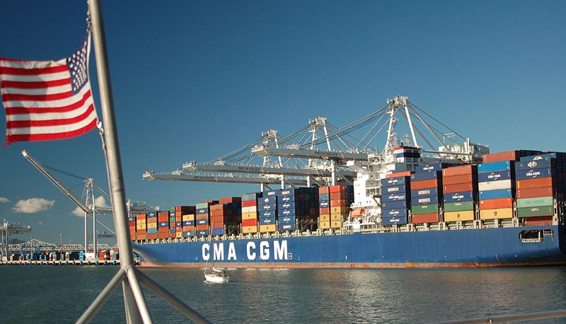 An image of a cargo ship transporting multiple colorful shipping containers. An American flag waves in the foreground.